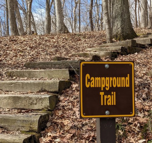 A wooden sign reading "Campground Trail" next to a set of steps leading through a wooded area with fallen leaves.
