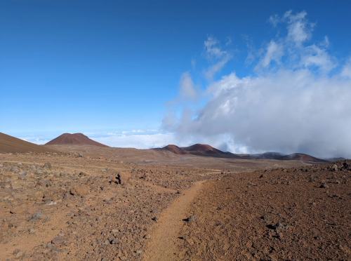 A rocky landscape with a dirt path leading towards distant volcanic mountains under a clear blue sky with clouds.