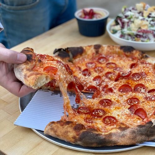 A hand reaches for a slice of pepperoni pizza, with melted cheese stretching from the pie on a metal tray.