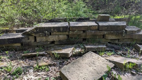 A weathered stone wall partially collapsed, surrounded by greenery and scattered stones on the ground.