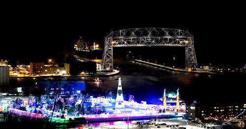 Aerial view of a festive waterfront with colorful lights, a tall Christmas tree, and a bridge illuminated at night.