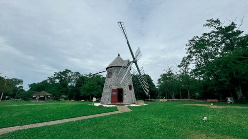 A wooden windmill stands in a grassy area surrounded by trees under a cloudy sky.