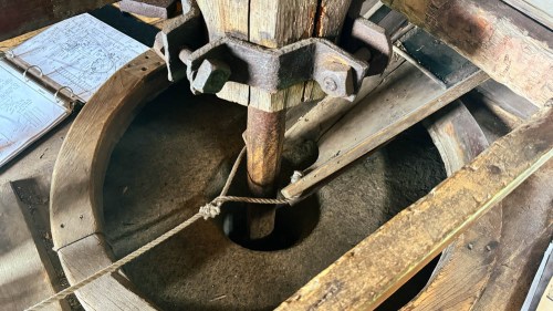 Close-up of a wooden millstone with a metal shaft, surrounded by rustic wooden beams and a blueprint in the background.