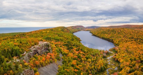 A long lake is surrounded by forested bluffs with vibrant autumn foliage under a gray sky with Lake Superior visible in the background.