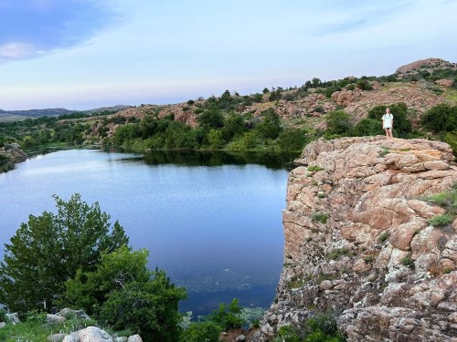 A person stands on a rocky ledge overlooking a calm lake surrounded by greenery and hills under a clear sky.