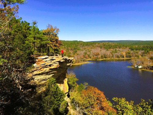 A person in a red shirt stands on a rocky cliff overlooking a serene lake surrounded by autumn foliage.
