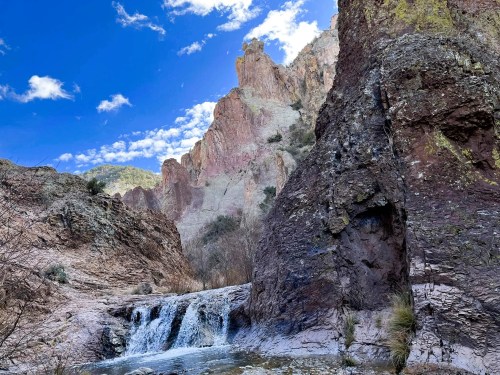 A serene landscape featuring a waterfall cascading over rocks, surrounded by towering cliffs and a blue sky with clouds.