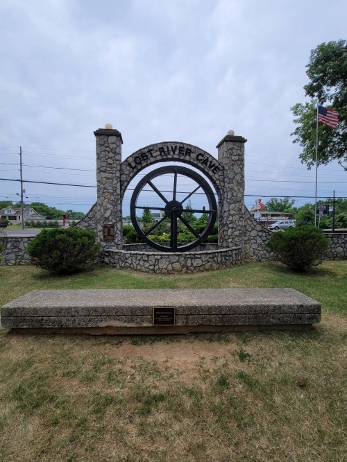 Stone structure with a large wheel, "Lost River Cave" sign, and a bench in front, surrounded by grass and trees.