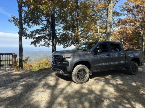 A gray pickup truck parked on a gravel overlook surrounded by trees with autumn foliage and a scenic view in the background.