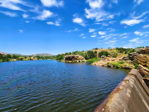 A serene lake surrounded by rocky hills and lush greenery under a bright blue sky with scattered clouds.