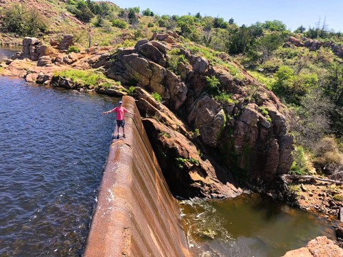 A person stands on a rocky dam, overlooking a calm body of water surrounded by lush greenery and rugged terrain.