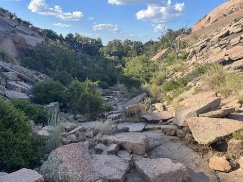Rocky landscape with scattered boulders, greenery, and a clear blue sky with fluffy clouds.