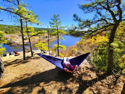 A person relaxes in a hammock overlooking a serene lake surrounded by trees and autumn foliage.