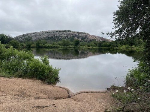 A serene landscape featuring a calm lake reflecting a rocky hill, surrounded by lush greenery under a cloudy sky.