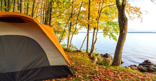 A camping tent set up beside a lakeshore in autumn, surrounded by trees with bright yellow and green leaves. Fallen leaves cover the ground, and calm blue water stretches out beyond the rocky shoreline under a lightly clouded sky.
