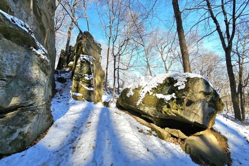 Snow-covered rocks and trees under a clear blue sky, with shadows cast on a snowy path.