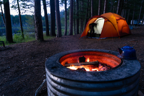A campsite at dusk with a glowing campfire in the foreground and an orange tent set up nearby.