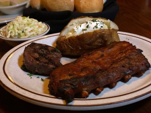 A plate featuring ribs, a steak, a baked potato with sour cream, and a side of coleslaw.