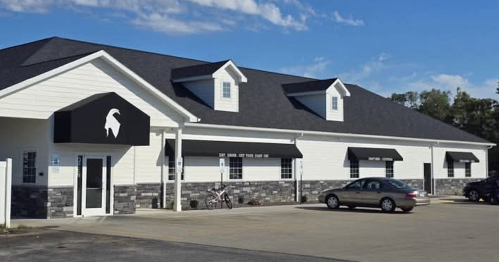 A low white building has black awnings over its windows, and the black awning above its door depicts the head of a goat.
