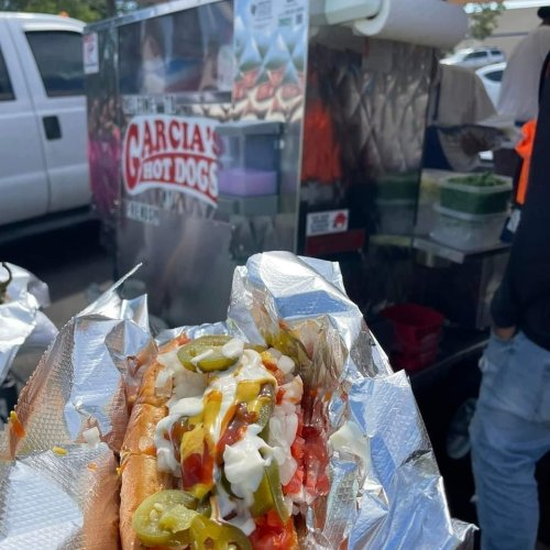 A close-up of a loaded hot dog with toppings, held in front of a food cart labeled "Garcia's Hot Dogs."
