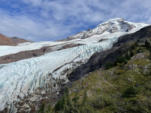A glacier flows down a rocky mountain, surrounded by green vegetation and a blue sky with scattered clouds.