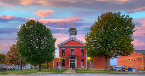 Historic red-brick courthouse with white trim and a central cupola, framed by two large leafy trees at sunset. The sky is filled with pink and purple clouds, and warm lights illuminate the building’s entrance.