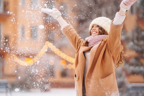 A joyful woman in a winter coat and hat throws snow in the air, smiling against a snowy backdrop with warm lights.