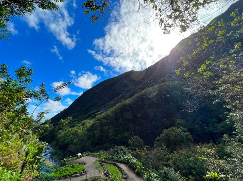 A scenic view of a mountain with lush greenery, a winding path, and a bright sky with clouds. Sunlight shines through.