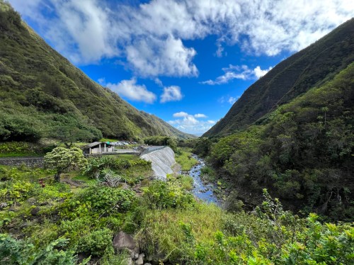 A lush valley with a river, surrounded by steep green hills and a blue sky with scattered clouds.