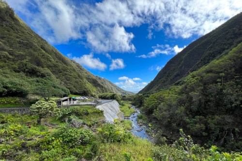 A lush valley with steep green mountains, a clear river, and a small building under a blue sky with scattered clouds.