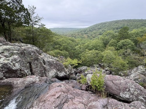 A rocky landscape with lush green trees and rolling hills under a cloudy sky. Water flows over the rocks in the foreground.