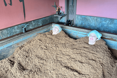 Two people relaxing in a spa, partially buried in warm, brown sand, with towels on their heads.