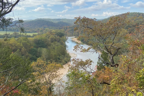 A scenic view of a winding river surrounded by trees and rolling hills, with autumn foliage under a partly cloudy sky.