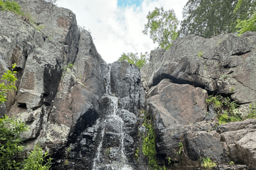 A small waterfall cascades over rocky cliffs, surrounded by greenery and a cloudy sky above.