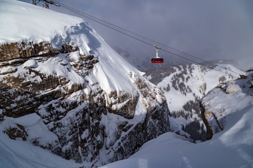 A red gondola glides over a snowy mountain landscape, with steep cliffs and a valley below.