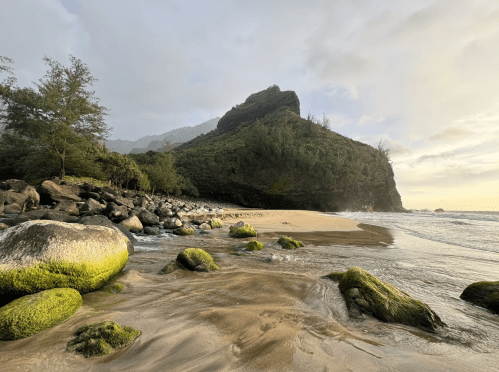 A serene beach scene with moss-covered rocks, gentle waves, and a lush green mountain in the background under a cloudy sky.