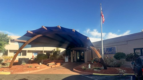 Entrance of a building with a large blue canopy, American flag, and desert landscaping. Clear blue sky above.