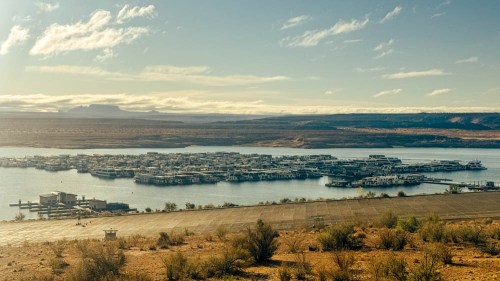 A scenic view of a marina with houseboats on a calm lake, surrounded by desert landscape and distant mountains.