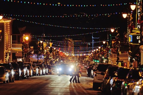 A festive street at night, adorned with colorful lights and decorations, with cars parked along the sides.