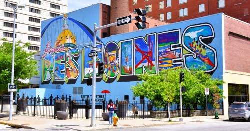A colorful mural showcasing the words 'Greater Des Moines' on the side of a building, featuring artistic scenes of the city.