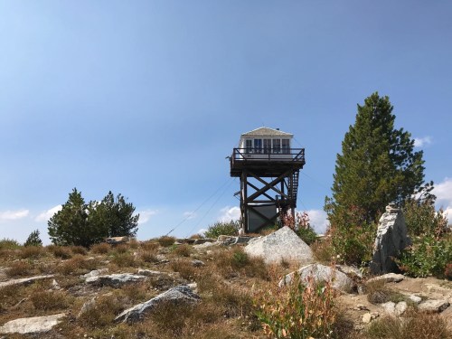 A wooden fire lookout tower stands on a rocky hillside, surrounded by sparse vegetation and under a clear blue sky.
