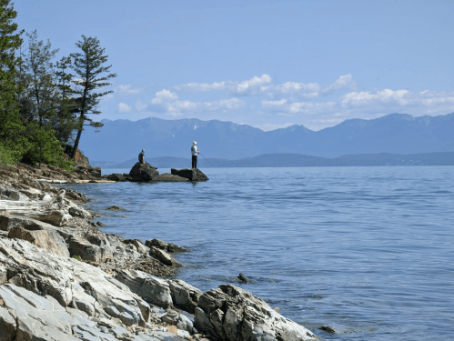 Two people fishing on rocky shore with mountains and clear blue sky in the background.