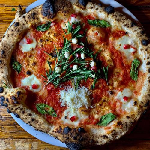 A freshly baked pizza topped with tomato sauce, mozzarella, fresh basil, rosemary, and pomegranate seeds on a wooden table.