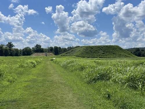 A grassy path leads to two large earthen mounds under a blue sky with fluffy clouds.
