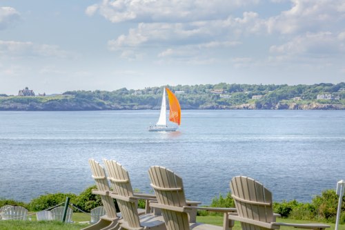 A sailboat with an orange sail glides across calm water, with Adirondack chairs in the foreground and greenery in the background.