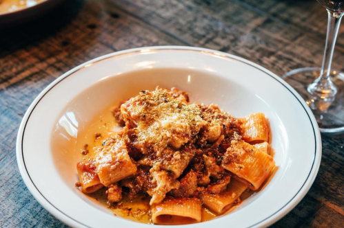 A plate of rigatoni pasta topped with a savory meat sauce and sprinkled with breadcrumbs, served on a wooden table.