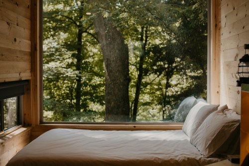 Cozy bedroom with a large window overlooking a lush green forest. Natural light fills the space.