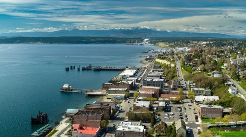 Aerial view of a coastal town with a waterfront, docks, and mountains in the background under a clear blue sky.