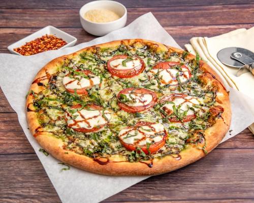 A freshly baked pizza topped with tomatoes, basil, and drizzled with sauce, served with cheese and red pepper flakes.