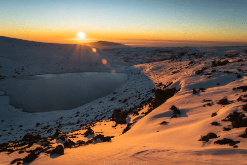 Sunrise over a snowy landscape with a frozen lake, casting warm light on the snow-covered terrain.